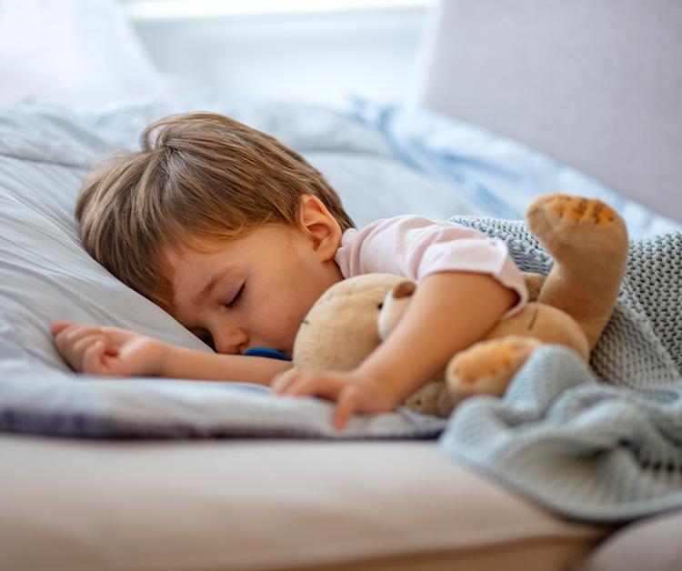 A young child sleeping peacefully on a bed, cuddling a stuffed teddy bear and covered with a soft blue blanket.