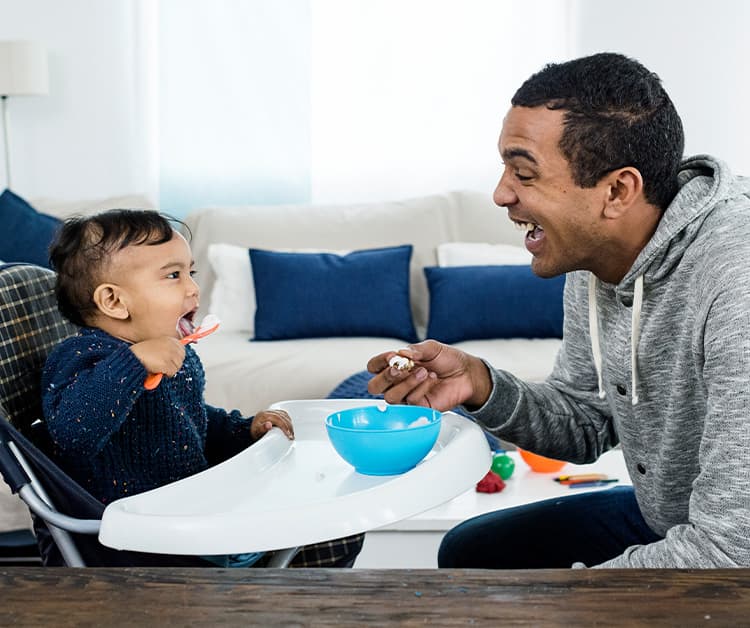 Toddler son in a high chair being fed yogurt by dad