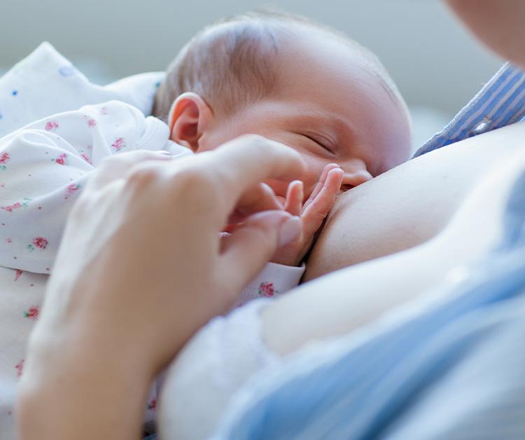 A newborn baby wearing a floral onesie breastfeeding, while gently holding onto the parent’s hand for comfort.