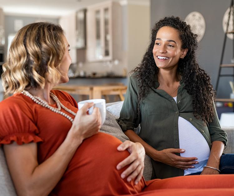 Two pregnant women sitting on the couch talking with one holding a cup of coffee